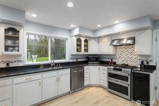 kitchen featuring wall chimney range hood, dark stone countertops, a sink, white cabinetry, and appliances with stainless steel finishes