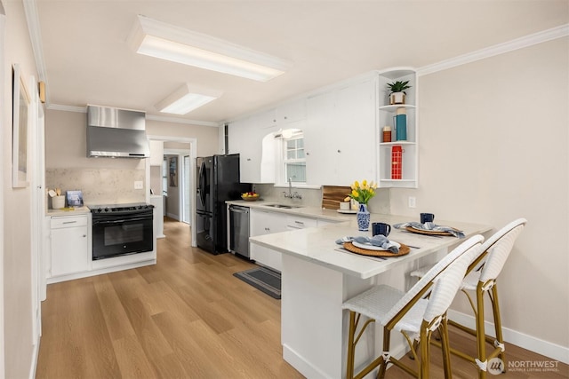 kitchen with wall chimney range hood, a peninsula, light countertops, white cabinetry, and black appliances