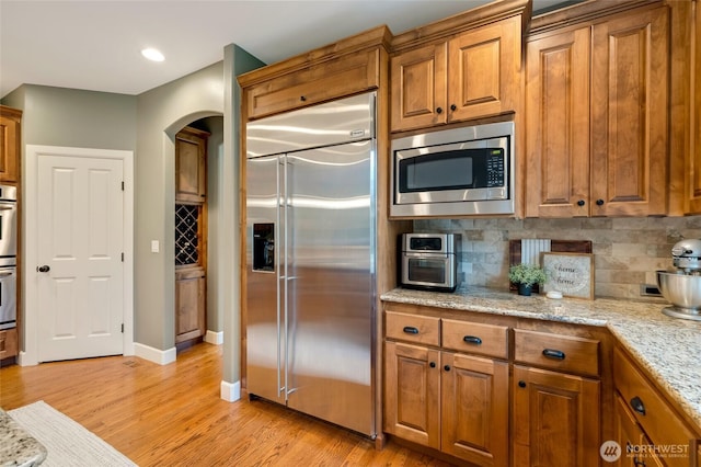 kitchen with tasteful backsplash, arched walkways, built in appliances, brown cabinetry, and light wood finished floors