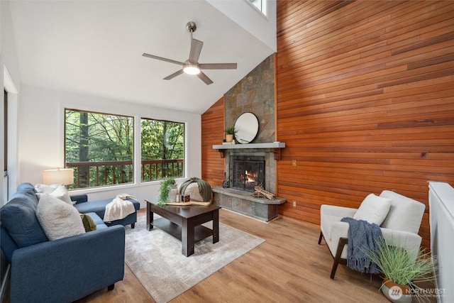 living room featuring a ceiling fan, a large fireplace, light wood-type flooring, high vaulted ceiling, and wooden walls