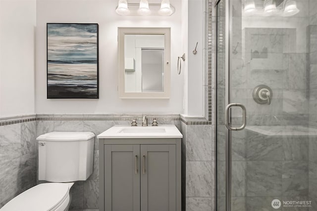 bathroom with a shower stall, vanity, toilet, wainscoting, and tile walls