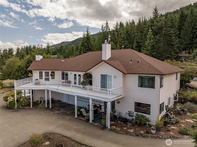 back of house featuring a chimney, a shingled roof, a forest view, and driveway