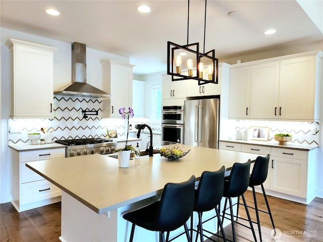 kitchen with wall chimney range hood, appliances with stainless steel finishes, an island with sink, white cabinetry, and tasteful backsplash
