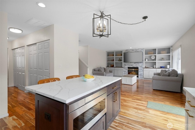 kitchen with an inviting chandelier, light wood-style floors, a fireplace, stainless steel microwave, and light stone counters