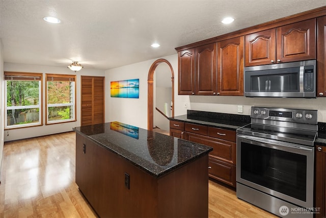 kitchen featuring arched walkways, appliances with stainless steel finishes, light wood-style floors, dark stone counters, and a kitchen island