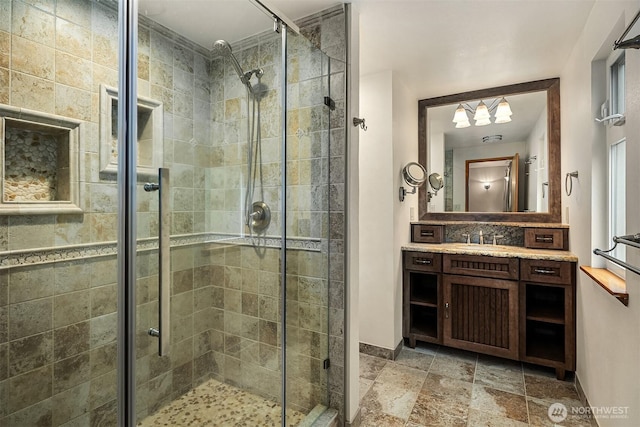bathroom featuring stone tile floors, vanity, a chandelier, a stall shower, and baseboards