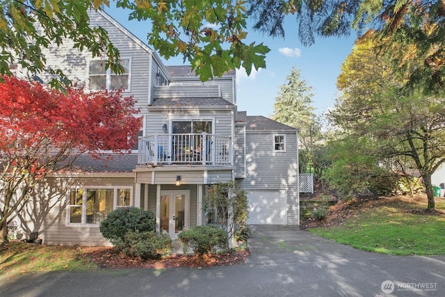 shingle-style home with a balcony, french doors, driveway, a garage, and a shingled roof