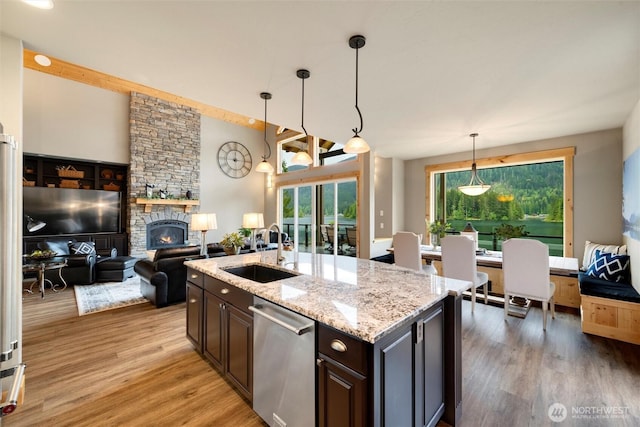 kitchen with a sink, stainless steel dishwasher, light wood-style flooring, a stone fireplace, and light stone counters