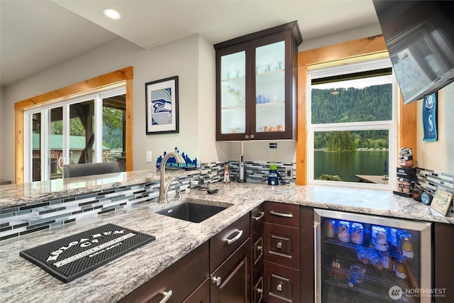 kitchen with wine cooler, a sink, dark brown cabinetry, light stone countertops, and glass insert cabinets