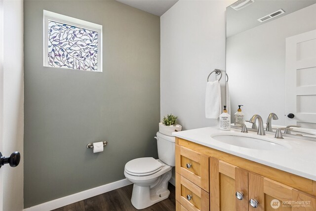 half bathroom with vanity, toilet, baseboards, wood finished floors, and visible vents