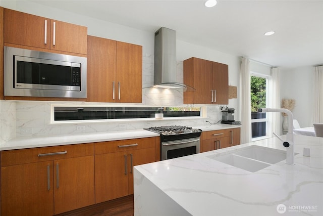 kitchen featuring a sink, wall chimney range hood, brown cabinets, light stone countertops, and appliances with stainless steel finishes
