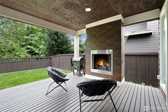 wooden terrace with a fenced backyard, a warm lit fireplace, a grill, and a yard