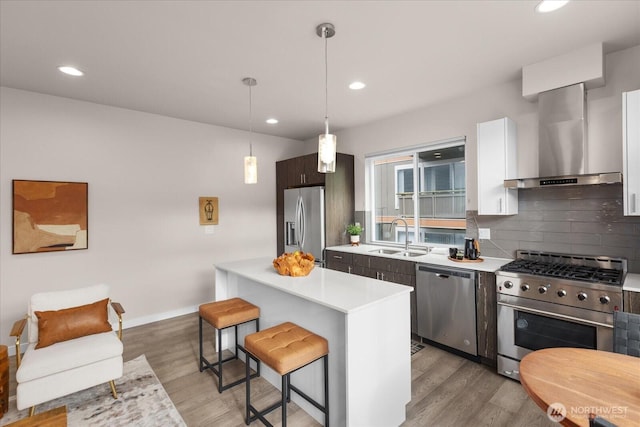 kitchen featuring wall chimney exhaust hood, appliances with stainless steel finishes, light countertops, a sink, and a breakfast bar area