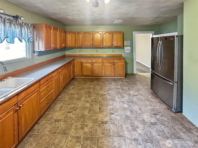 kitchen featuring stainless steel refrigerator with ice dispenser, a sink, light countertops, brown cabinets, and stone finish flooring