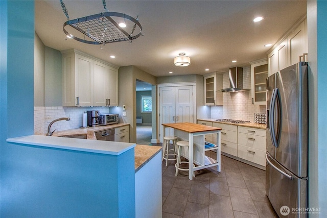 kitchen with glass insert cabinets, wall chimney range hood, appliances with stainless steel finishes, a peninsula, and a toaster