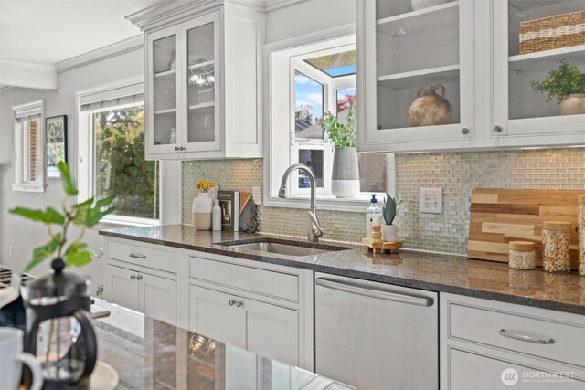 kitchen featuring dark stone counters, stainless steel dishwasher, a sink, decorative backsplash, and ornamental molding
