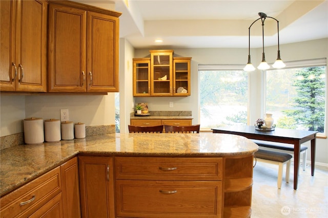 kitchen featuring a peninsula, glass insert cabinets, light stone countertops, brown cabinets, and pendant lighting