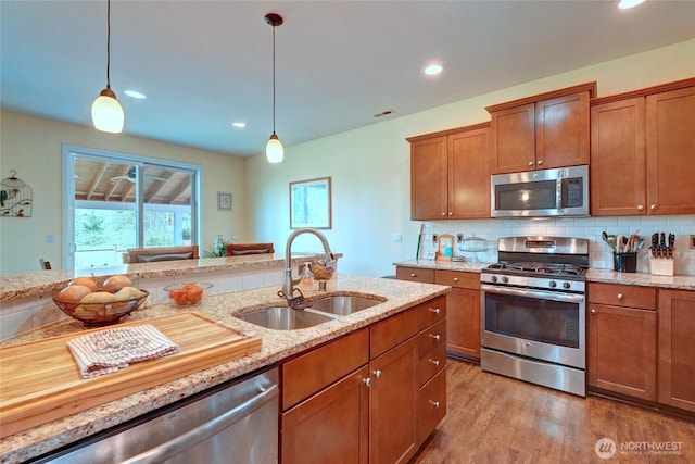 kitchen with appliances with stainless steel finishes, a sink, brown cabinetry, hanging light fixtures, and wood finished floors