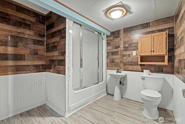 bathroom featuring wood finished floors, combined bath / shower with glass door, wood walls, and toilet