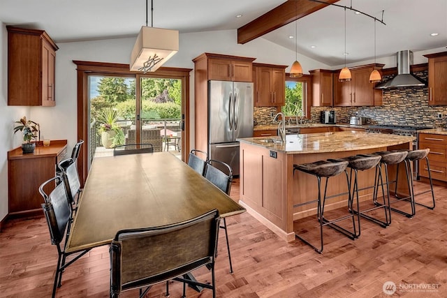 kitchen with wall chimney exhaust hood, lofted ceiling with beams, a center island with sink, decorative backsplash, and stainless steel refrigerator