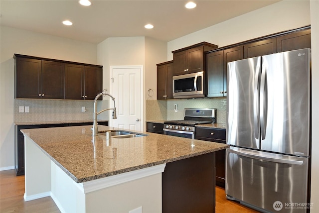 kitchen with stainless steel appliances, a sink, dark brown cabinets, light stone counters, and an island with sink