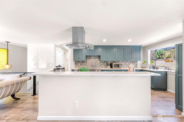 kitchen featuring island exhaust hood, light countertops, stainless steel microwave, light wood-style floors, and decorative backsplash