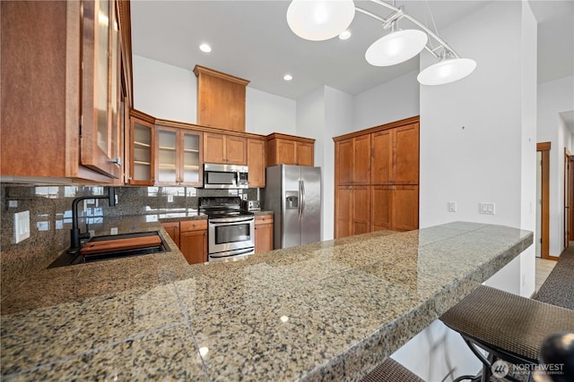 kitchen featuring a sink, backsplash, glass insert cabinets, brown cabinets, and appliances with stainless steel finishes