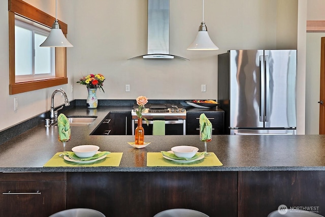 kitchen featuring a sink, dark countertops, wall chimney range hood, appliances with stainless steel finishes, and hanging light fixtures