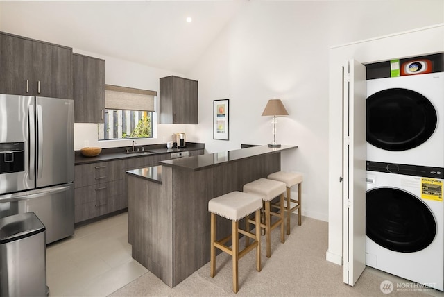 kitchen with a sink, stainless steel refrigerator with ice dispenser, a breakfast bar area, stacked washer and clothes dryer, and dark countertops