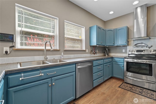 kitchen with blue cabinets, wall chimney range hood, appliances with stainless steel finishes, and light wood-type flooring