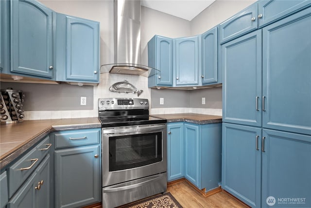 kitchen featuring blue cabinets, wall chimney exhaust hood, dark countertops, light wood-style floors, and electric stove