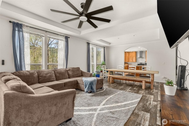 living room featuring a tray ceiling, dark wood-type flooring, arched walkways, a ceiling fan, and baseboards