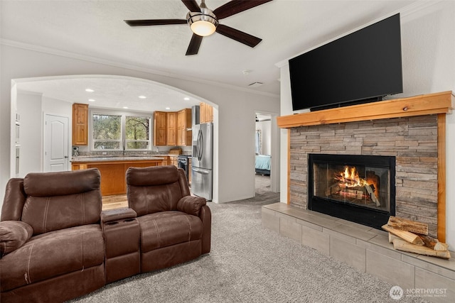 living area featuring crown molding, arched walkways, a ceiling fan, a stone fireplace, and light carpet
