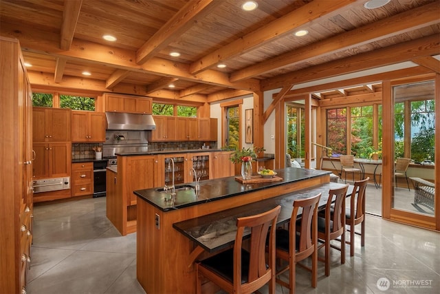 kitchen featuring under cabinet range hood, a center island with sink, wood ceiling with exposed beams, dark countertops, and backsplash