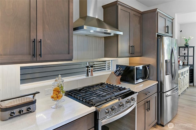 kitchen with wall chimney range hood, dark brown cabinets, light stone counters, light wood finished floors, and stainless steel appliances