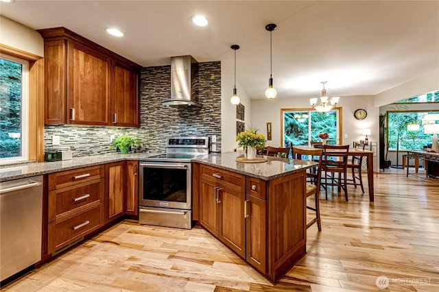 kitchen with wall chimney range hood, brown cabinetry, appliances with stainless steel finishes, and a peninsula
