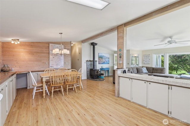 kitchen with white cabinets, vaulted ceiling with beams, ceiling fan with notable chandelier, wood walls, and a wood stove