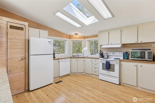 kitchen featuring light wood finished floors, visible vents, vaulted ceiling with skylight, white appliances, and wooden walls