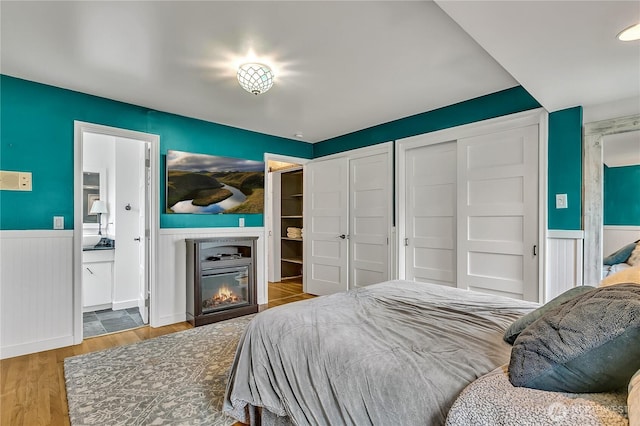 bedroom featuring a glass covered fireplace, wood finished floors, two closets, connected bathroom, and a wainscoted wall