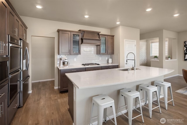 kitchen with a kitchen bar, a sink, appliances with stainless steel finishes, premium range hood, and dark wood-style flooring