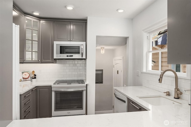 kitchen featuring glass insert cabinets, a sink, backsplash, gray cabinetry, and stainless steel appliances