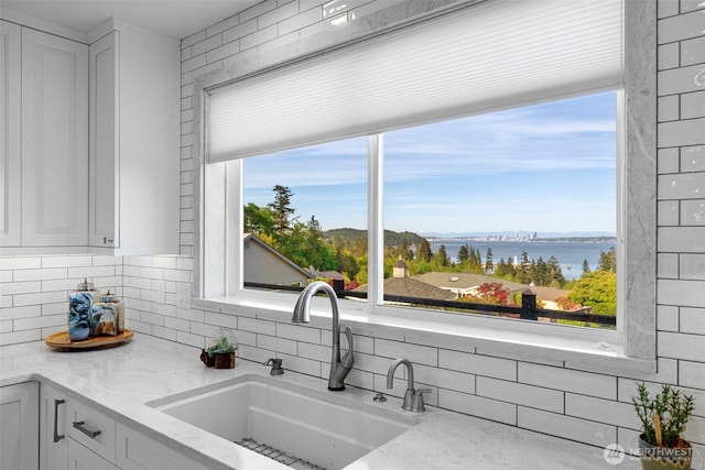 kitchen featuring a sink, backsplash, a water view, light stone countertops, and white cabinetry