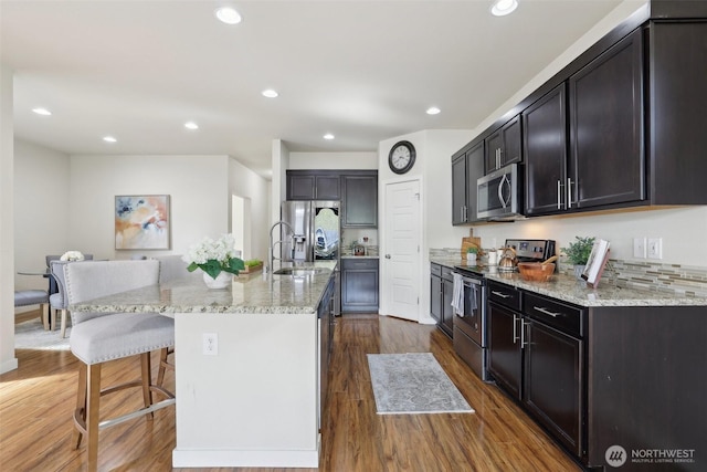 kitchen with stainless steel appliances, a kitchen island with sink, dark wood-style flooring, a kitchen bar, and light stone countertops