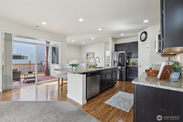 kitchen featuring a sink, stainless steel appliances, a center island with sink, light stone countertops, and dark wood-style floors