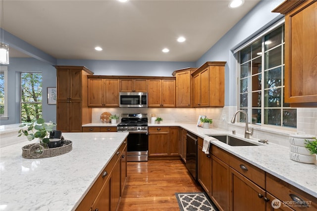 kitchen with appliances with stainless steel finishes, a sink, brown cabinets, wood finished floors, and light stone counters