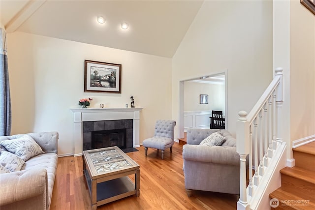 living room featuring high vaulted ceiling, a tiled fireplace, stairway, and light wood-type flooring