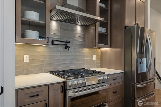 kitchen featuring stainless steel appliances, wall chimney range hood, glass insert cabinets, light stone countertops, and tasteful backsplash