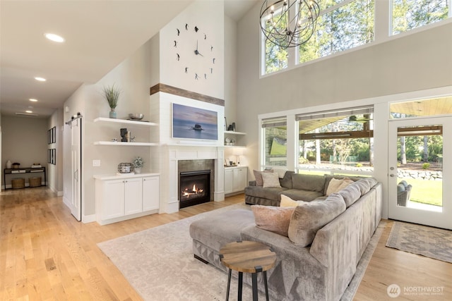 living area with a barn door, a glass covered fireplace, light wood-type flooring, a towering ceiling, and recessed lighting