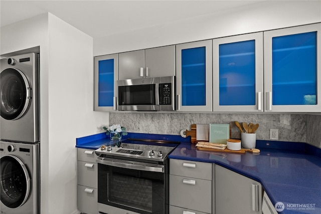 kitchen with glass insert cabinets, stacked washer and clothes dryer, dark countertops, stainless steel appliances, and tasteful backsplash