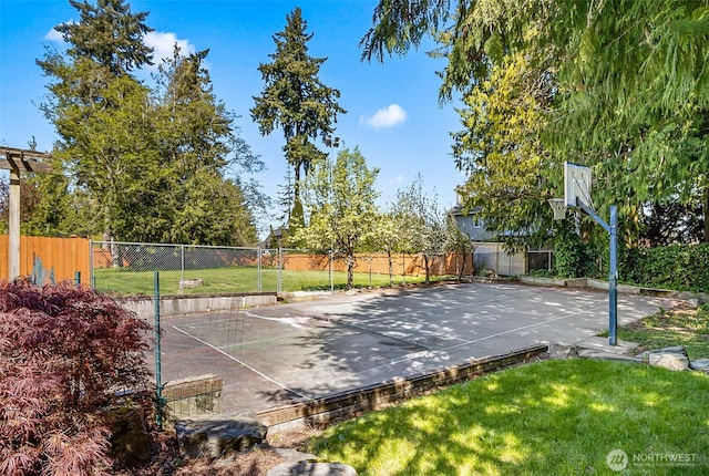 view of basketball court featuring a lawn, basketball hoop, and fence